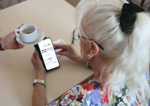 Einer Seniorin mit Handy in der Hand wird eine Tasse Kaffee gereicht